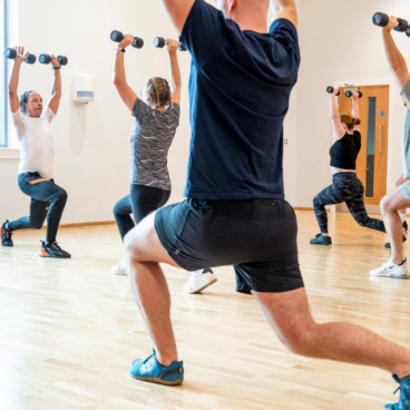 HOME Group of people performing single-leg squats while holding small dumbbells overhead in a fitness class